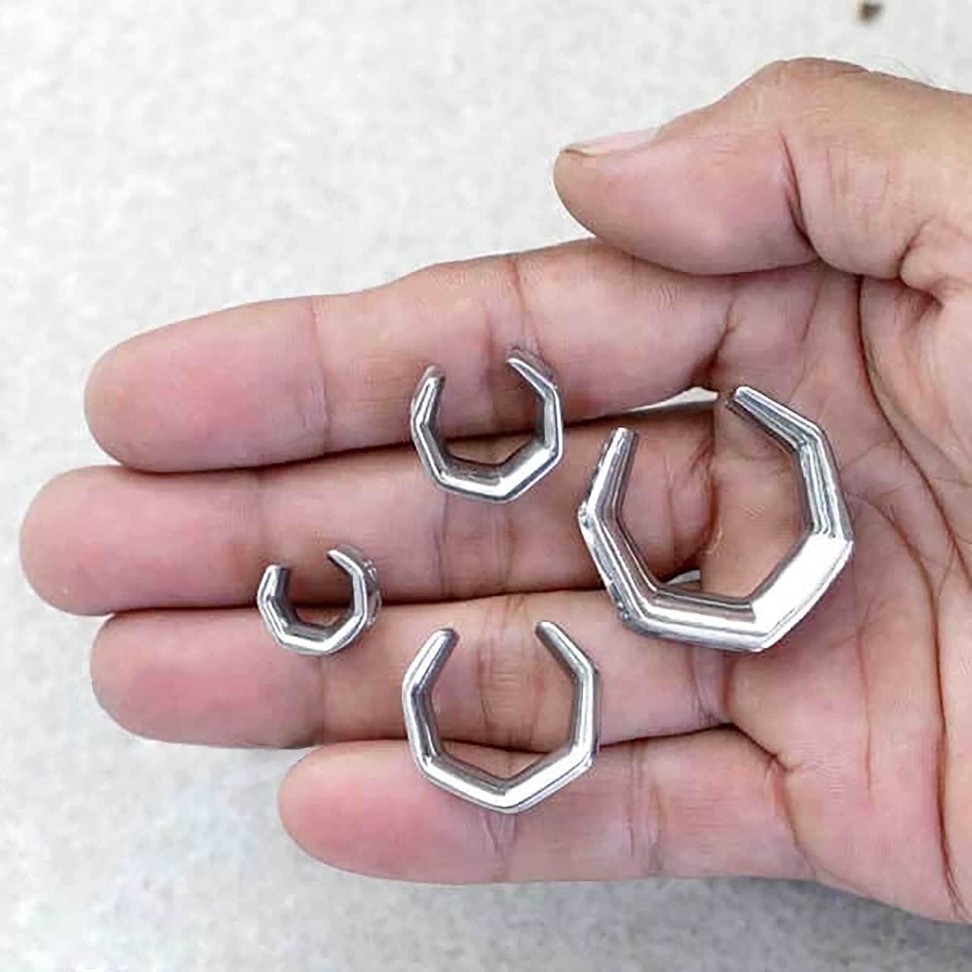 A hand is shown holding four small, hexagonal silver-colored objects of varying sizes, resembling tiny wrenches or open-ended hexagonal rings. These are the OCTAGON Minimalist Saddle Tunnels in Silver by Kaya Movement (ranging from 6mm to 25mm / 2g to 1 Inch), made from surgical steel gauges. The background is a textured, light-colored surface.